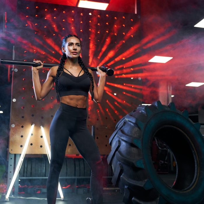 Front view of muscular woman in sportswear holding sledgehammer on shoulders near tire wheel. Full length portrait of female bodybuilder posing in gym, having rest after hard training, red lights.